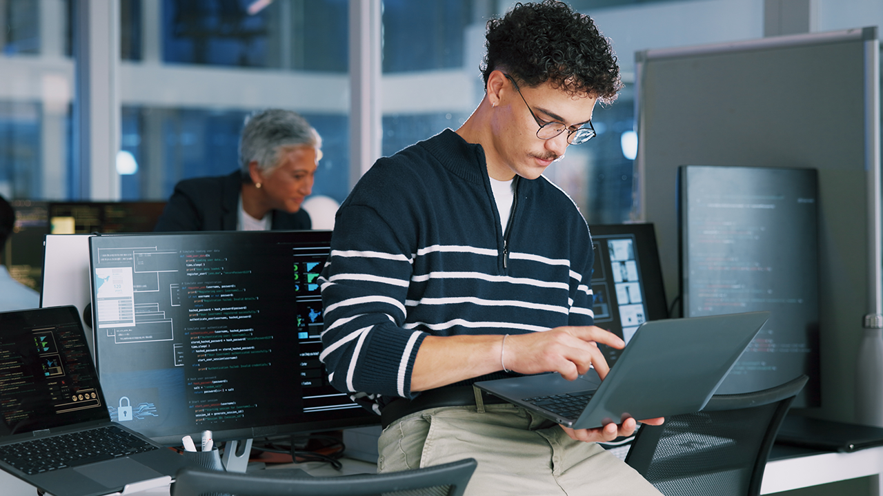 Man holding a laptop and facing multiple computer screens while improving code reliability through software testing techniques.