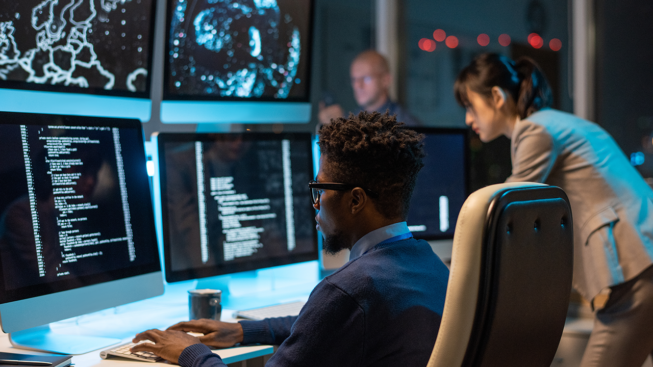 Man sitting at a desk analyzing computer screens while diagnosing and resolving operating system and network issues in a cybersecurity environment.